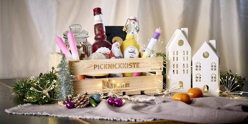 Auf einem Tisch steht eine Holzkiste. Daneben stehen verschiedene Dekoartikel, die mittels der Münsteraner Giebelhäuser und weihnachtlicher Dekoration ein festliche Stimmung schaffen. In der Kiste sind verschiedene Flaschen und Verpackungen mit Spezialitäten aus dem Münsterland zu sehen.