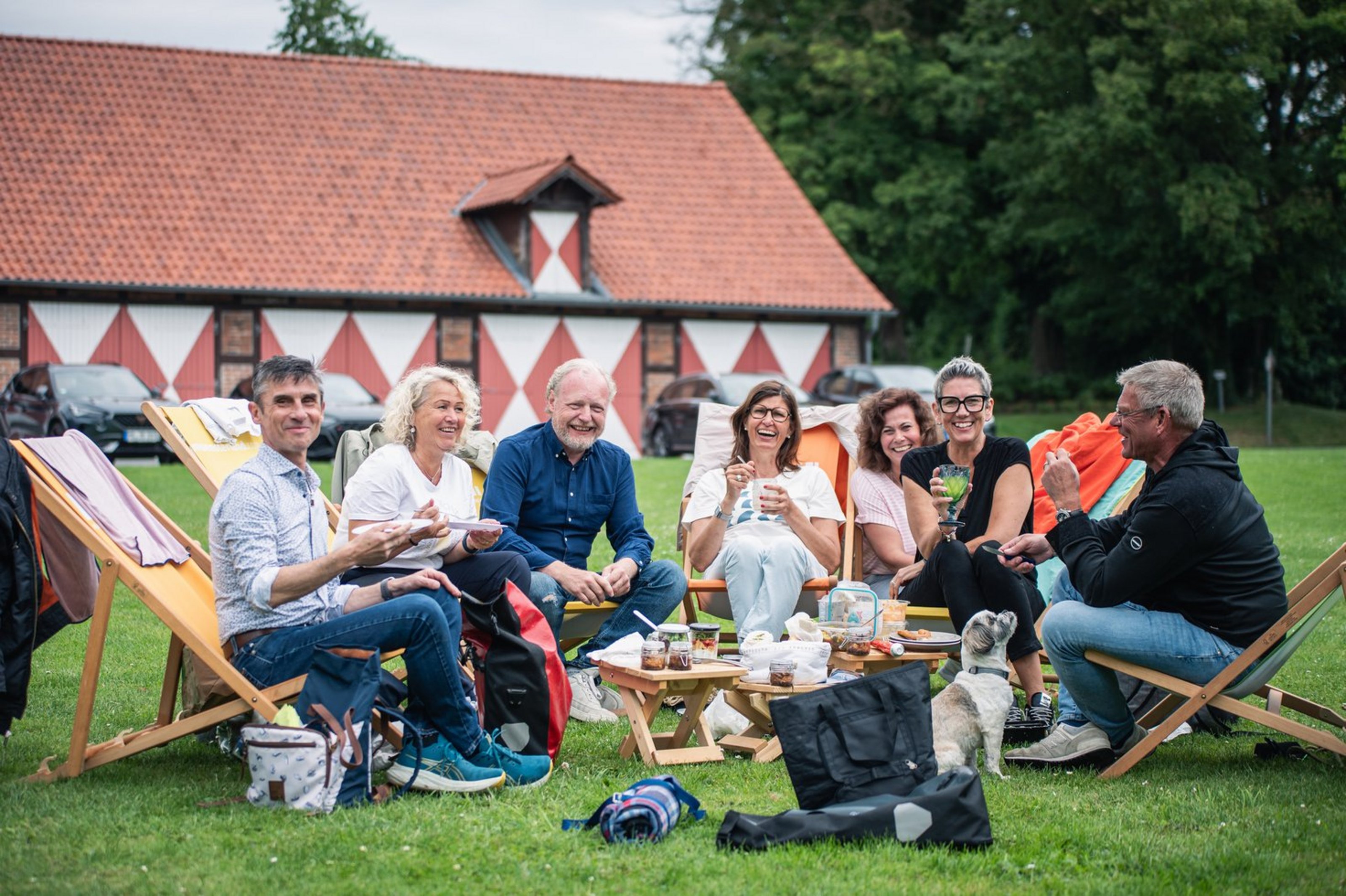Sieben Personen sitzen in Liegestühlen auf einer Wiese vor einem Fachwerk-Gebäude. Sie halten Teller oder Gläser in den Händen und in der Mitte stehen Hocker mit Dosen, Essen und Gläsern.