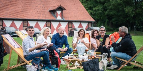 Sieben Personen sitzen in Liegestühlen auf einer Wiese vor einem Fachwerk-Gebäude. Sie halten Teller oder Gläser in den Händen und in der Mitte stehen Hocker mit Dosen, Essen und Gläsern.