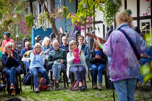 Eine Person mit Gitarre steht vor einem sitzenden Publikum, das in die Hände klatscht. Die Szene spielt sich auf einem gepflastertem Hof ab, im Hintergrund steht ein Fachwerkhaus.