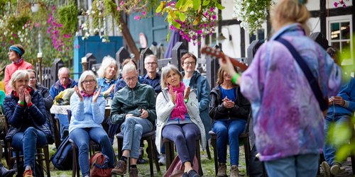 Eine Person mit Gitarre steht vor einem sitzenden Publikum, das in die Hände klatscht. Die Szene spielt sich auf einem gepflastertem Hof ab, im Hintergrund steht ein Fachwerkhaus.