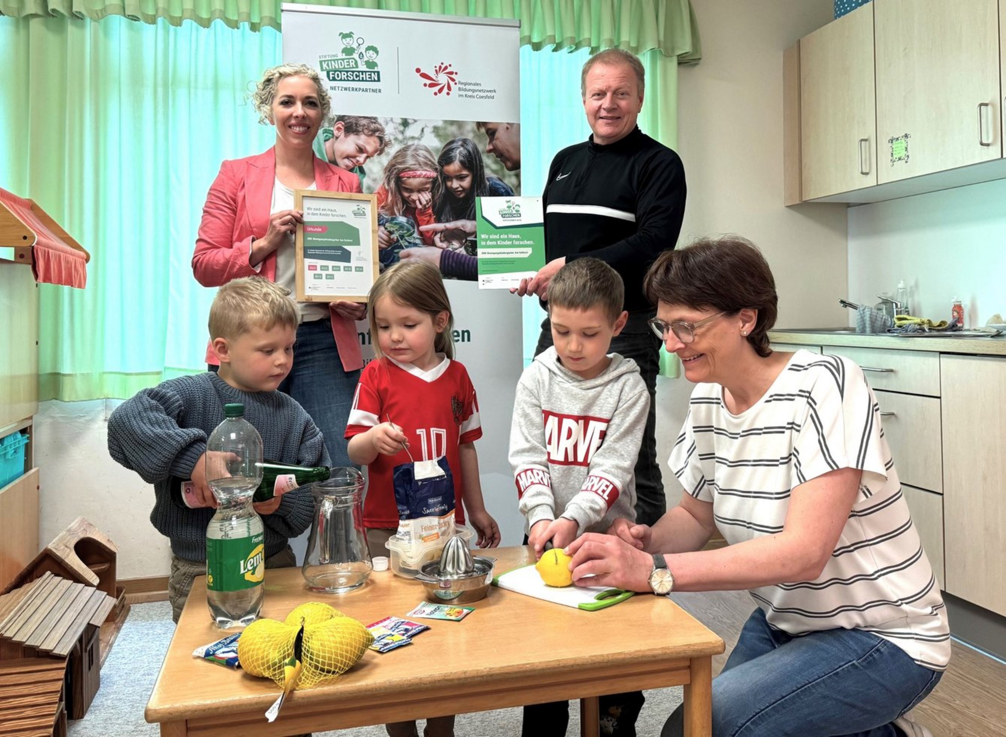 Drei Kinder experimentieren mit einer Erzieherin an einem Tisch, zwei Erwachsene im Hintergrund halten eine Auszeichnungsurkunde. Der DRK-Kindergarten „Am Schloss“ in Senden wurde für MINT-Bildung erneut zertifiziert.