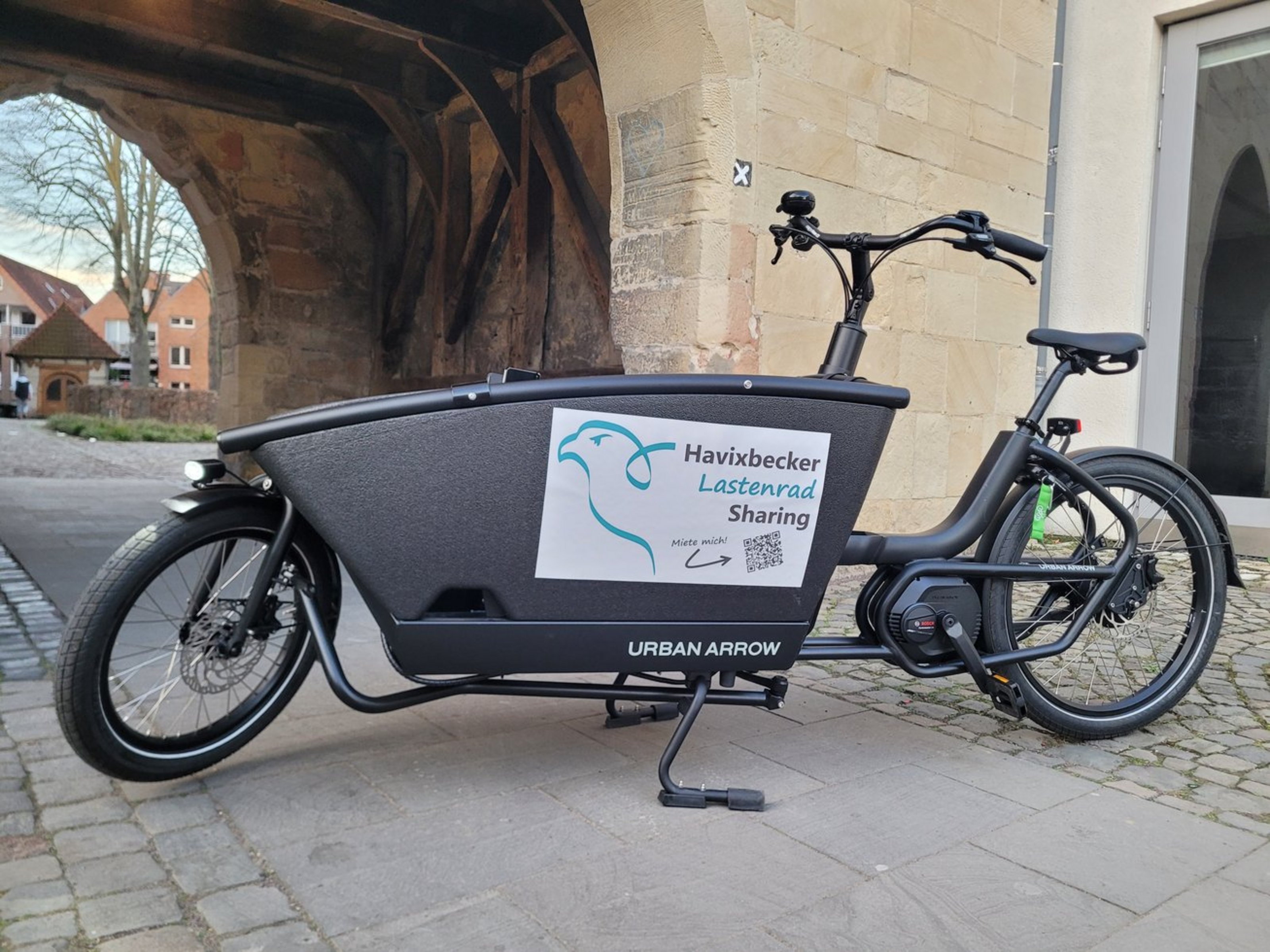 Das E-Lastenrad-Sharing ist im Frühling in vielen Kommunen im Kreis Coesfeld in die Saison gestartet (Foto: Gemeinde Havixbeck). Ein schwarzes E-Lastenrad ist vor einem hellen Steintor geparkt. Auf dem Lastenrad steht "Havixbecker Lastenrad Sharing". Darunter befindet sich ein QR-Code und daneben der Umriss eines Vogels. Hinter dem Tor sind ein Baum und Wohnhäuser zu sehen.