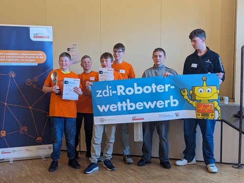 Eine Gruppe von sechs Jungen steht in einem Raum und posiert für ein Foto bei einem Robotikwettbewerb. Vier der Jungen tragen orangefarbene T-Shirts mit dem Aufdruck „Firebots“, zwei weitere tragen Freizeitkleidung. Einer der Jungen hält einen Pokal und eine Urkunde in den Händen. Drei weitere halten ebenfalls Urkunden. Zwei Jungen halten gemeinsam ein großes, blaues Banner mit der Aufschrift „zdi-Roboterwettbewerb“ und einer Zeichnung eines gelben Roboters. Im Hintergrund steht ein Aufsteller mit Informationen zum zdi-Netzwerk im Kreis Coesfeld in Nordrhein-Westfalen.