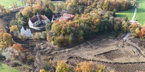Luftaufnahme der Burg Vischering, umgeben von herbstlichen Bäumen, mit sichtbaren Erdarbeiten und freigelegten Bereichen in der Außengräfte.