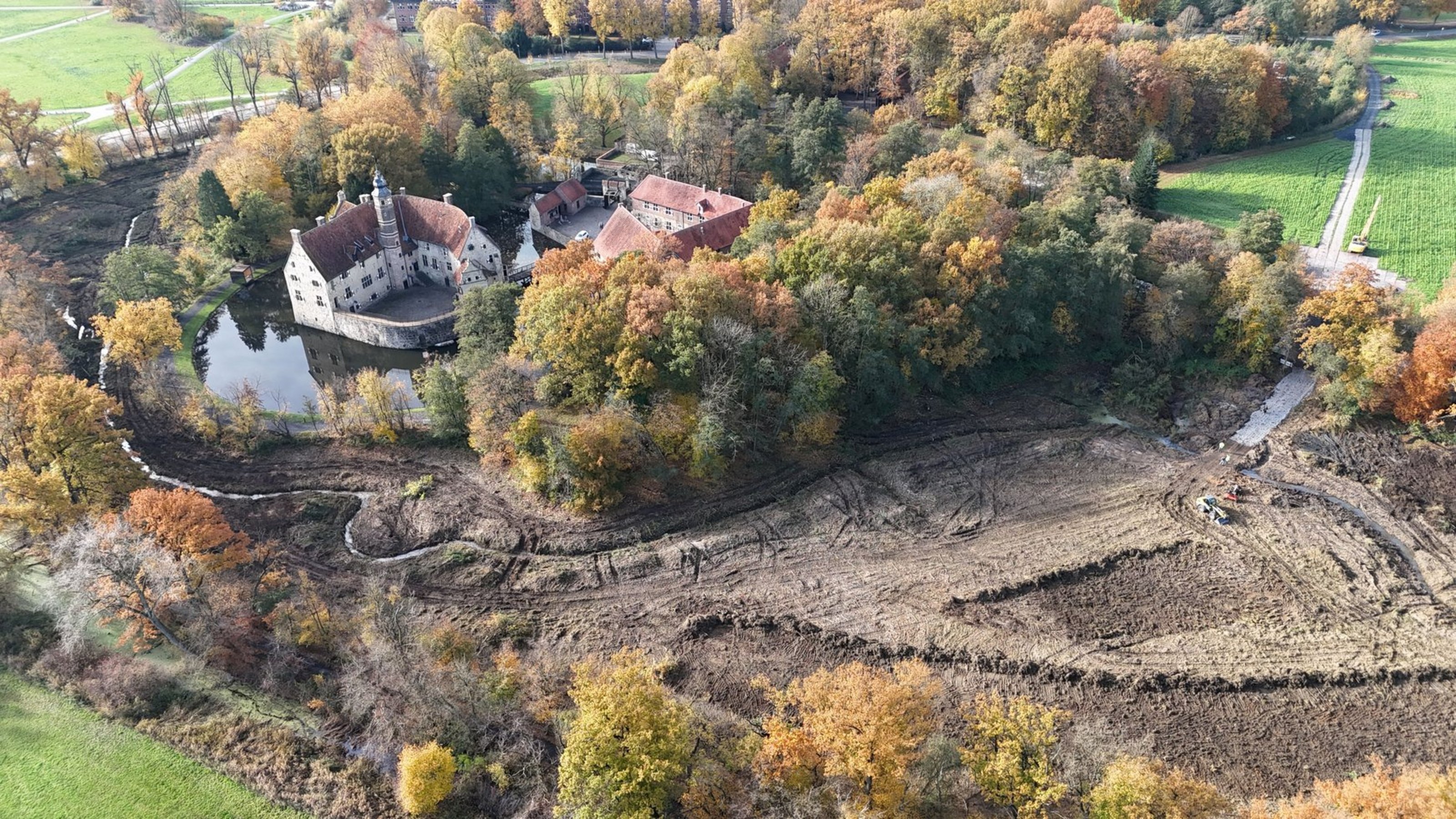 Luftaufnahme der Burg Vischering, umgeben von herbstlichen Bäumen, mit sichtbaren Erdarbeiten und freigelegten Bereichen in der Außengräfte.