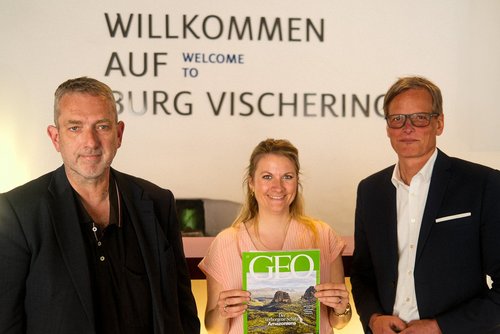 Drei Personen stehen nebeneinander vor einer Wand mit der Aufschrift „WILLKOMMEN AUF BURG VISCHERING“. In der Mitte hält eine Frau lächelnd ein GEO-Magazin mit grünem Titelblatt in den Händen. Links von ihr steht ein Mann in dunkler Kleidung mit grauen Haaren, rechts ein weiterer Mann in einem dunklen Anzug und weißem Hemd mit Brille.