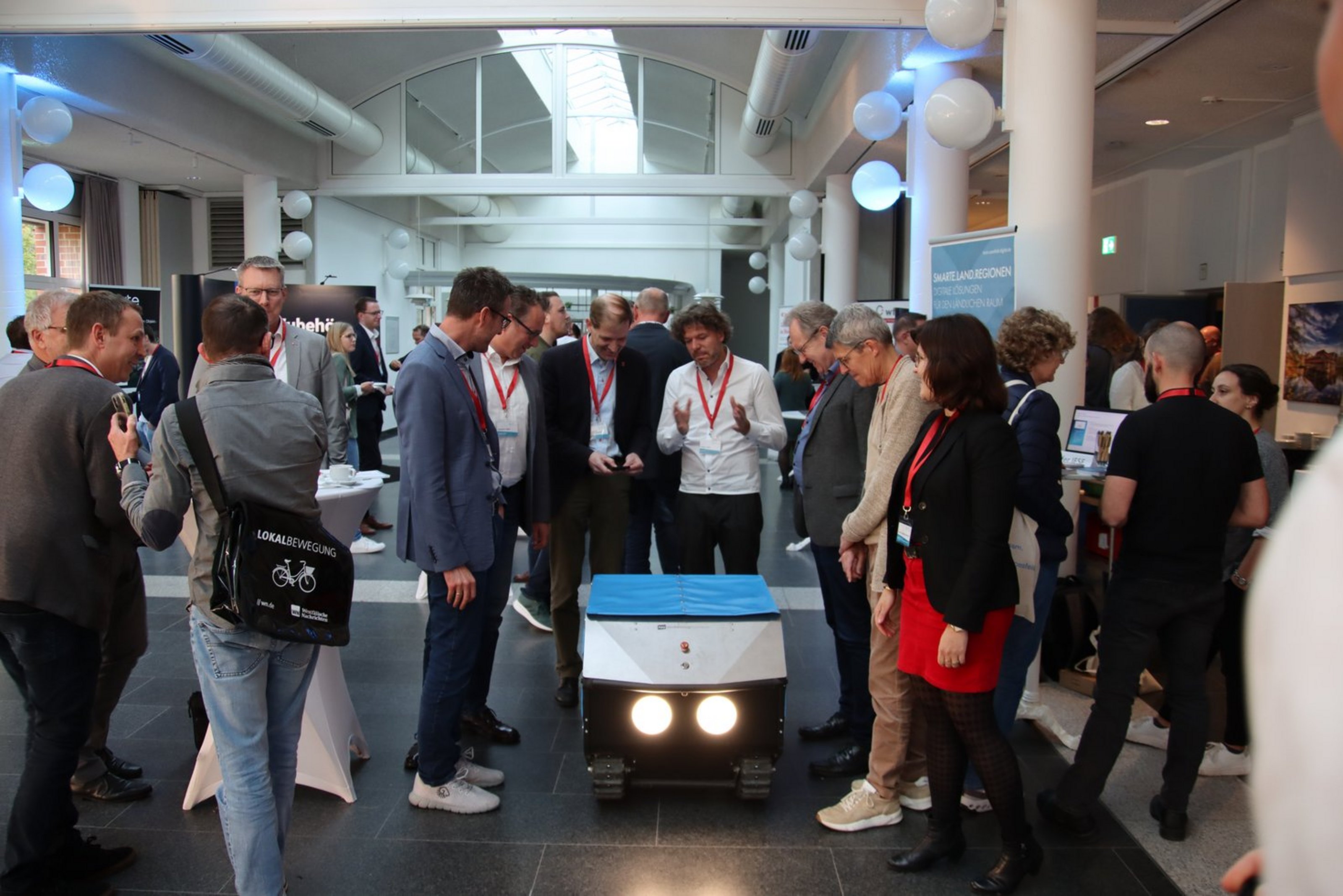 Teilnehmende einer Digitalisierungsveranstaltung stehen in Gruppen zusammen und tauschen sich an einem Ausstellungsstand aus. Im Mittelpunkt steht ein technisches Exponat, das von mehreren Personen betrachtet wird.