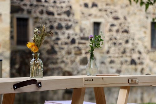 Zwei kleine Glasvasen mit schlichten Blumenarrangements stehen auf einer hellen Holzbank im Freien. Die linke Vase enthält getrocknete gelbe Blüten, Gräser und Ähren, während die rechte Vase frische grüne Zweige mit einer kleinen lila Blüte zeigt. Im Hintergrund ist eine grob gemauerte Steinwand mit kleinen Fenstern zu sehen.