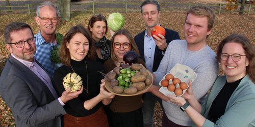 Acht Personen stehen in einer Gruppe und halten Gemüse und Eier nach oben in die Kamera. Sie stehen auf einem belaubten Untergrund, hinten befinden sich Parkplätze.