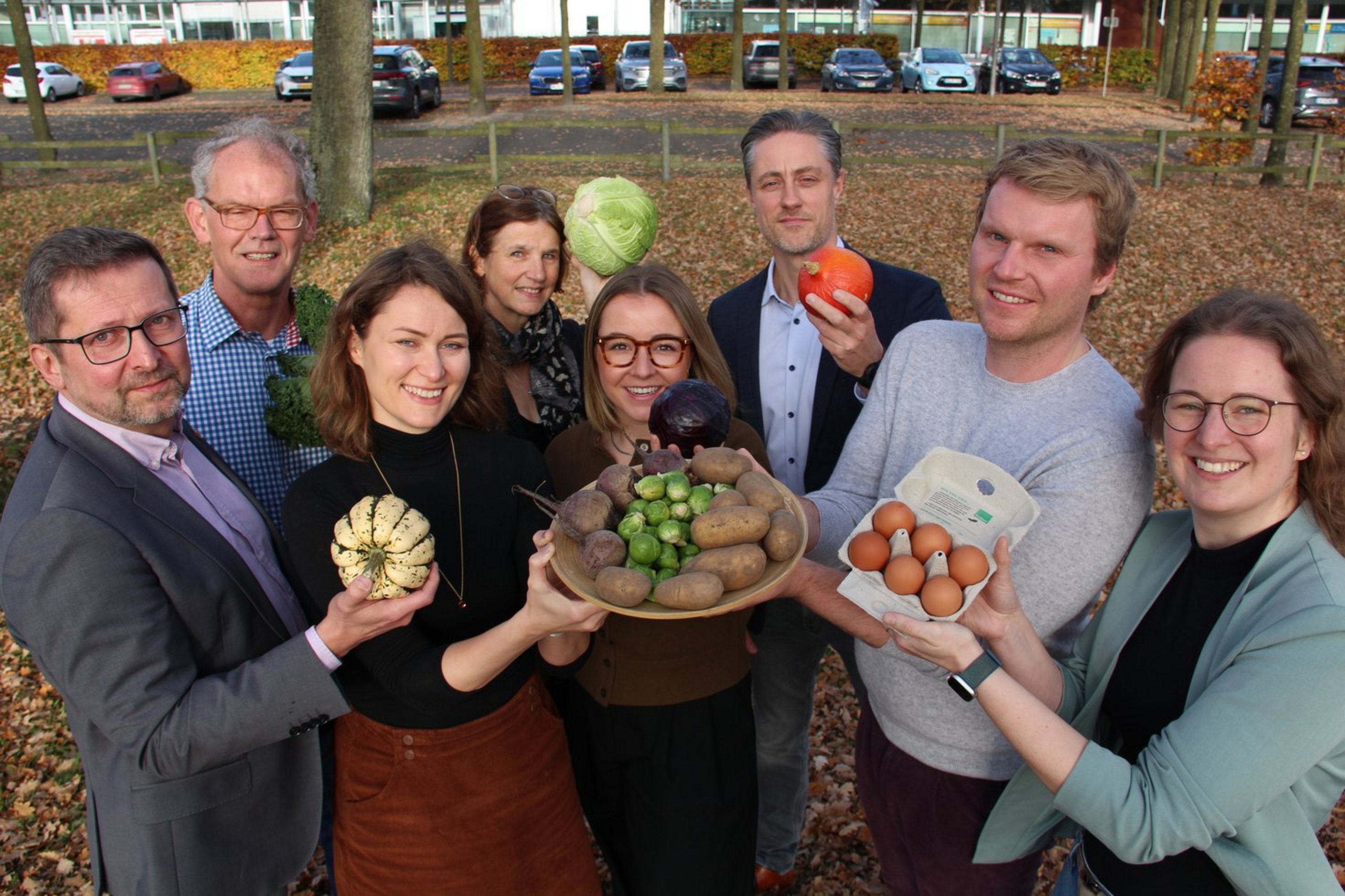 Acht Personen stehen in einer Gruppe und halten Gemüse und Eier nach oben in die Kamera. Sie stehen auf einem belaubten Untergrund, hinten befinden sich Parkplätze.