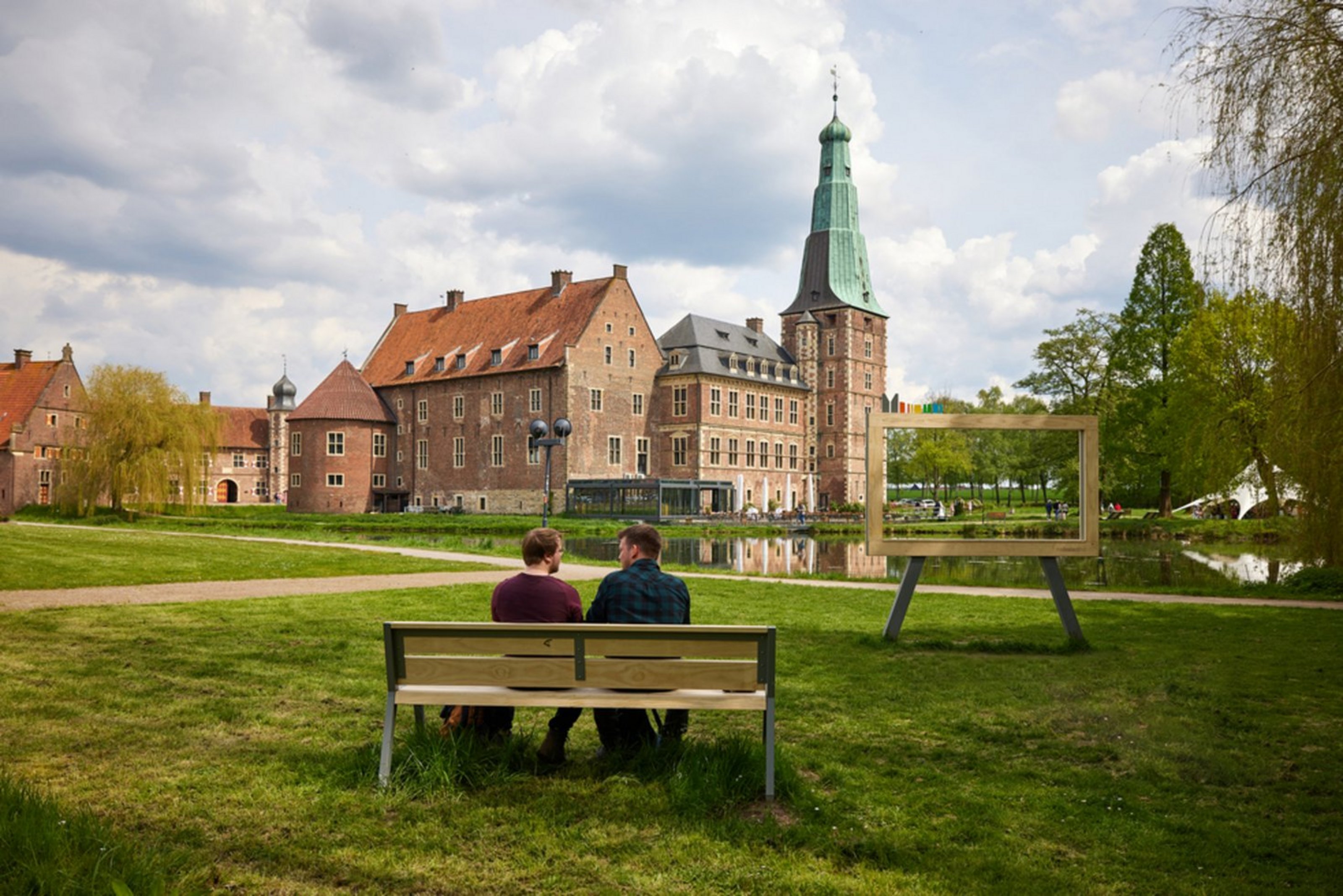 Ein Schloss aus rotem Backstein mit Wassergraben vor einer grünen Wiese ist zu sehen. Auf der Wiese sitzen zwei Personen auf einer Bank und schauen auf das Schloss.