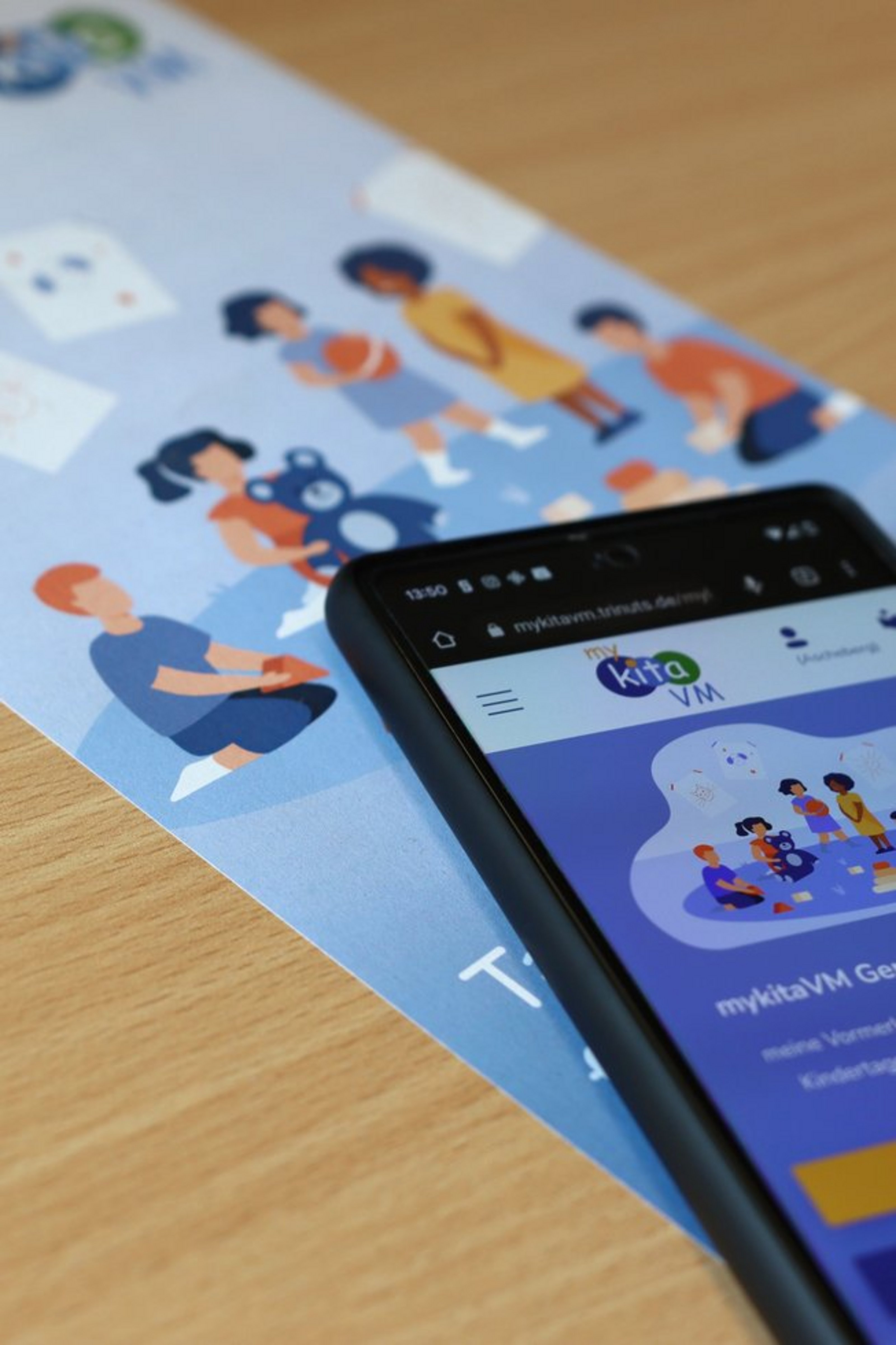 Ein Smartphone liegt auf einem Flyer mit einer Illustration von spielenden Kindern. Auf dem Smartphone ist die Webseite des Elternportals mykitaVM geöffnet.