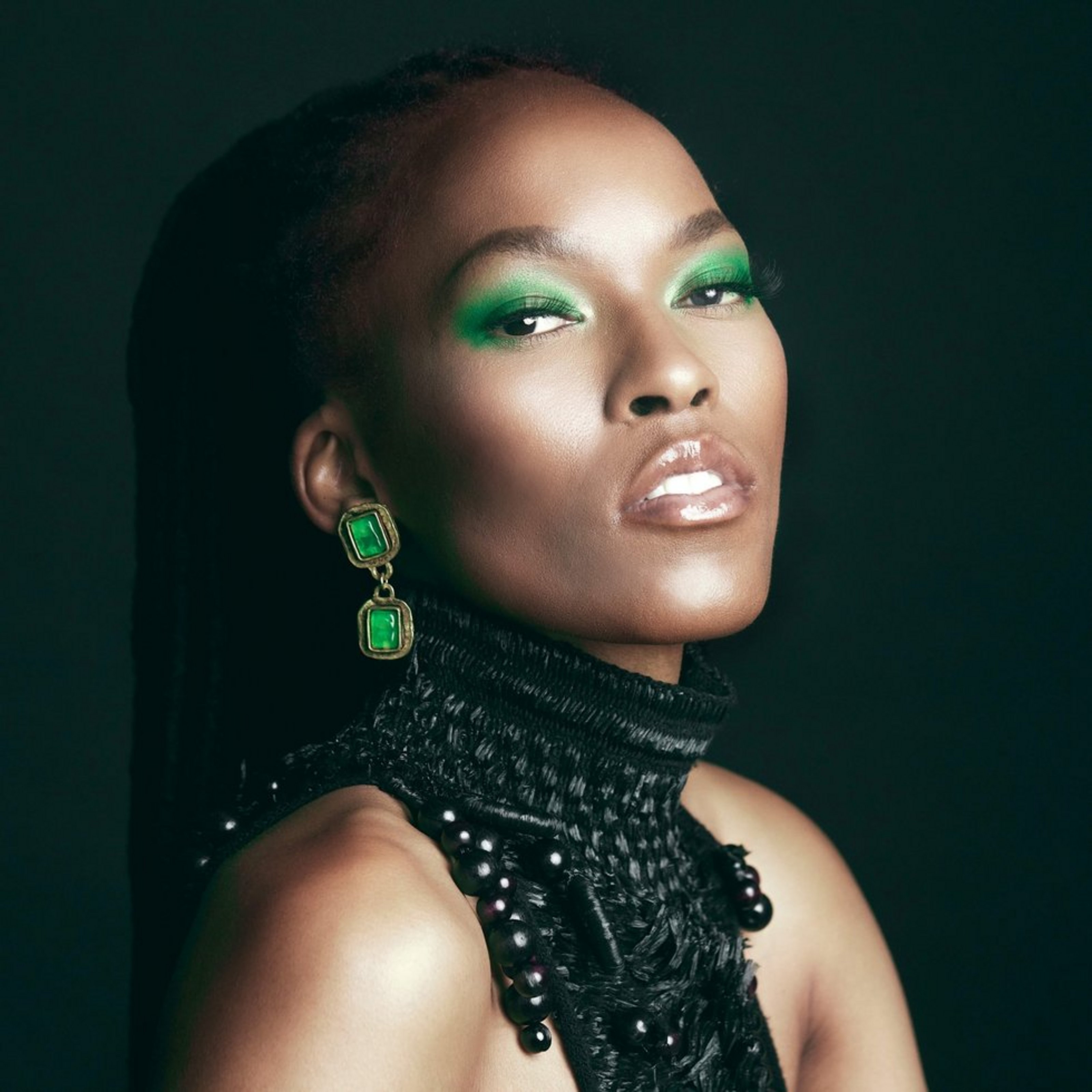 Das Foto ist eine Porträtaufnahme einer Frau mit grünem Lidschatten, grünen Ohrringen und schwarzem Oberteil.