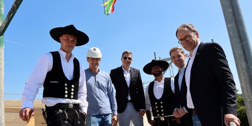 Sechs Männer stehen auf einem Baugerüst. Zwei von ihnen tragen die traditionelle Bekleidung von Zimmerleuten. Über ihnen hängt ein Gesteck mit bunten Schnüren.