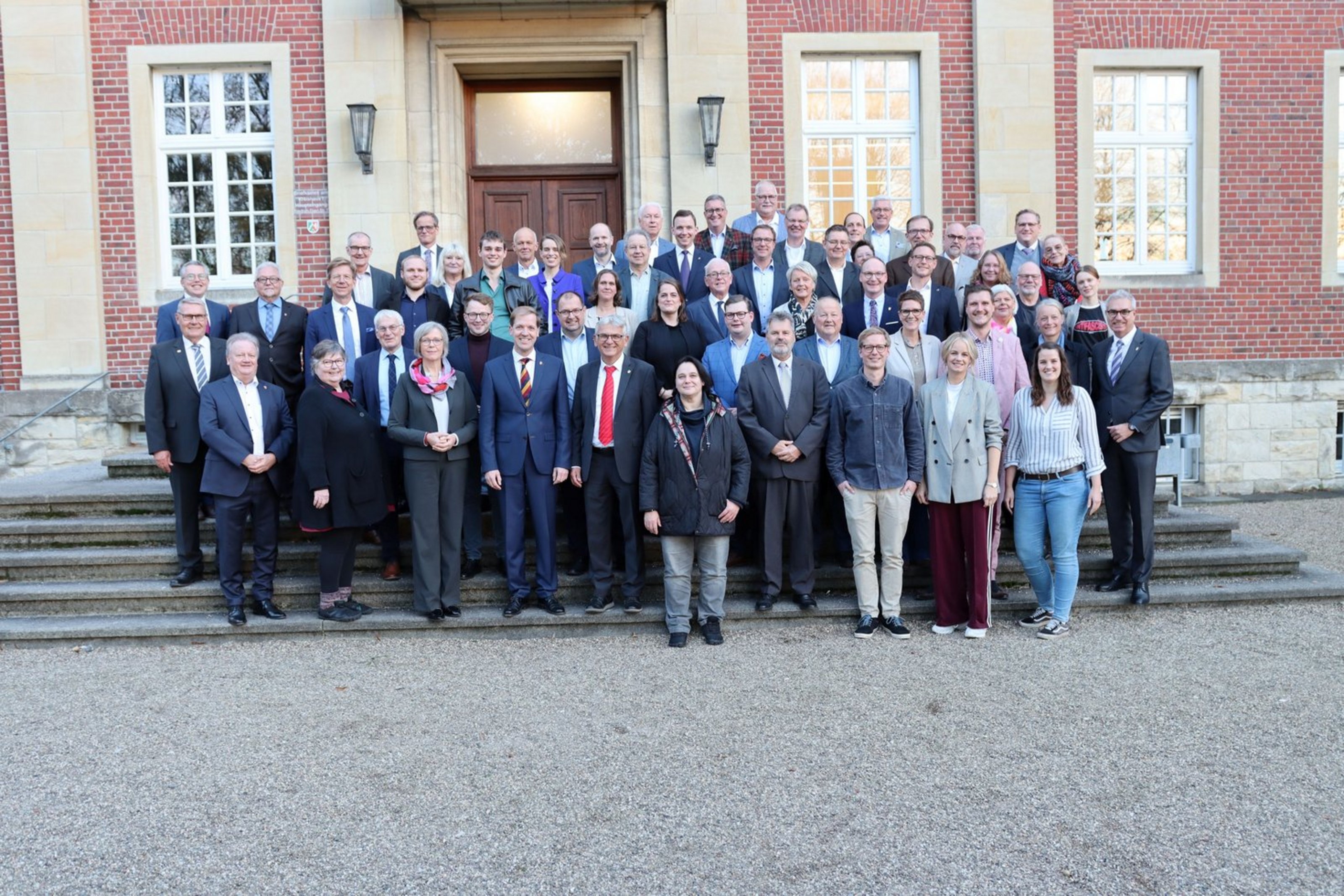 Eine Gruppe von ca.. 60 Personen steht auf den Treppenstufen vor einem alten Backsteingebäude in mehreren reihen hintereinander aufgereiht und schaut in die Kamera.