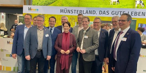 Zehn Männer und eine Frau stehen nebeneinander vor einer gelben Wand, auf der "Münsterland. Das gute Leben" steht.n 