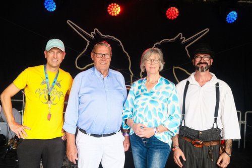 Drei Männer und eine Frau stehen vor einer schwarezn Wand, auf der zwei Einhornköpfe als Umrisse zu sehen sind. Einer der Personen trägt ein mittelalterliches Kostüm, die anderen tragen Bluse, Hemd oder Tshirt.