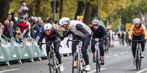 Vier Personen sind bei einem Radrennen zu sehen. Im Hintergrund stehen Zuschauer an der Straße.
