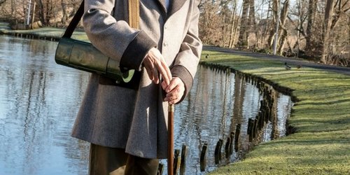 Ein Mann in Frack und Zylinder steht draußen auf einer grünen Wiese vor einem See.