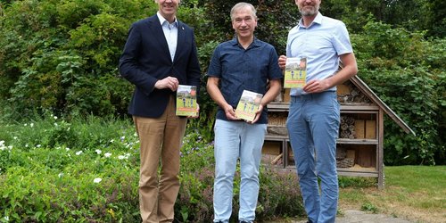 Drei Männer stehen vor einem Gebüsch. Sie halten kleine Hefte in der Hand und zeigen sie in die Kamera.