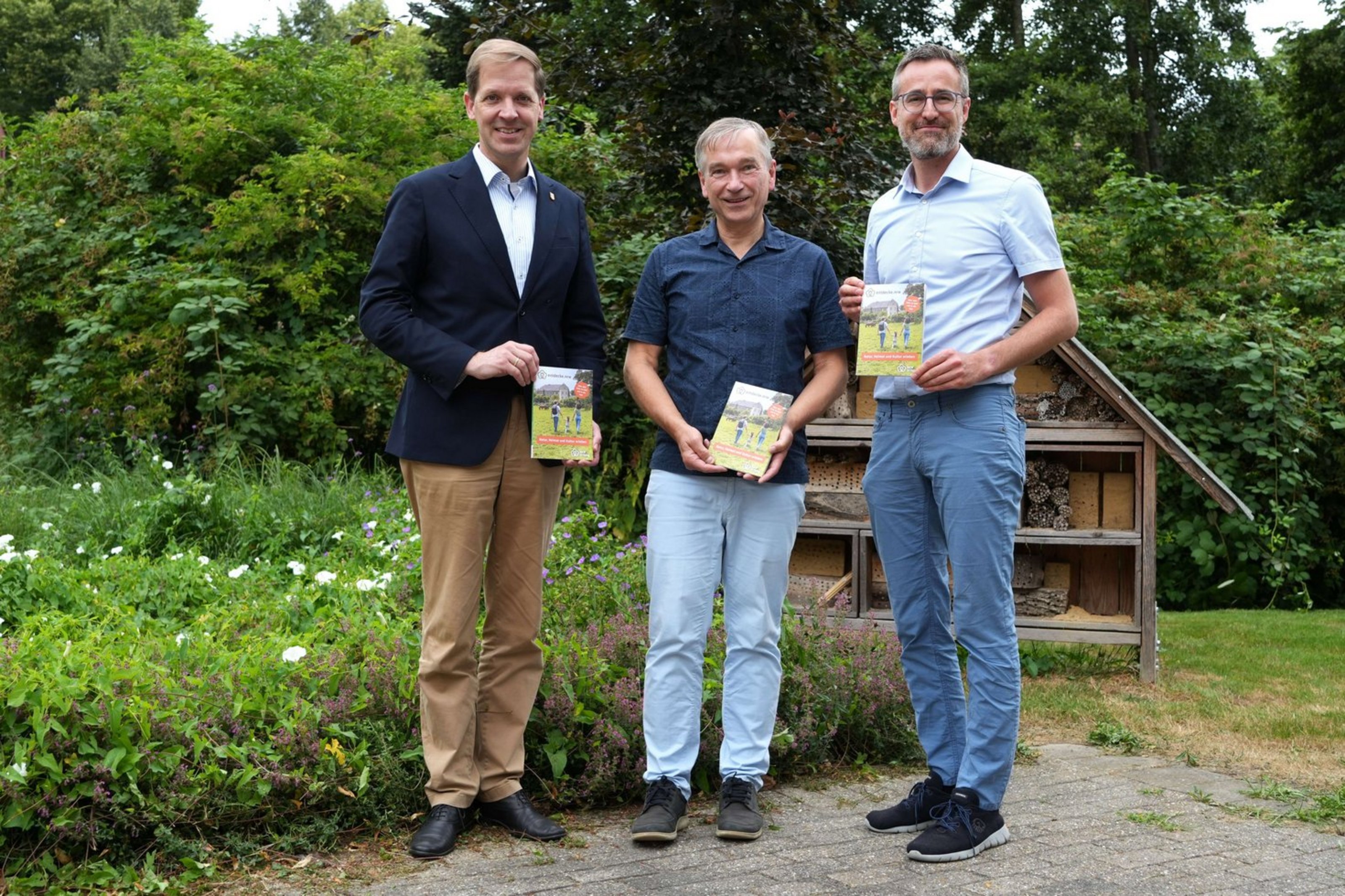 Drei Männer stehen vor einem Gebüsch. Sie halten kleine Hefte in der Hand und zeigen sie in die Kamera.