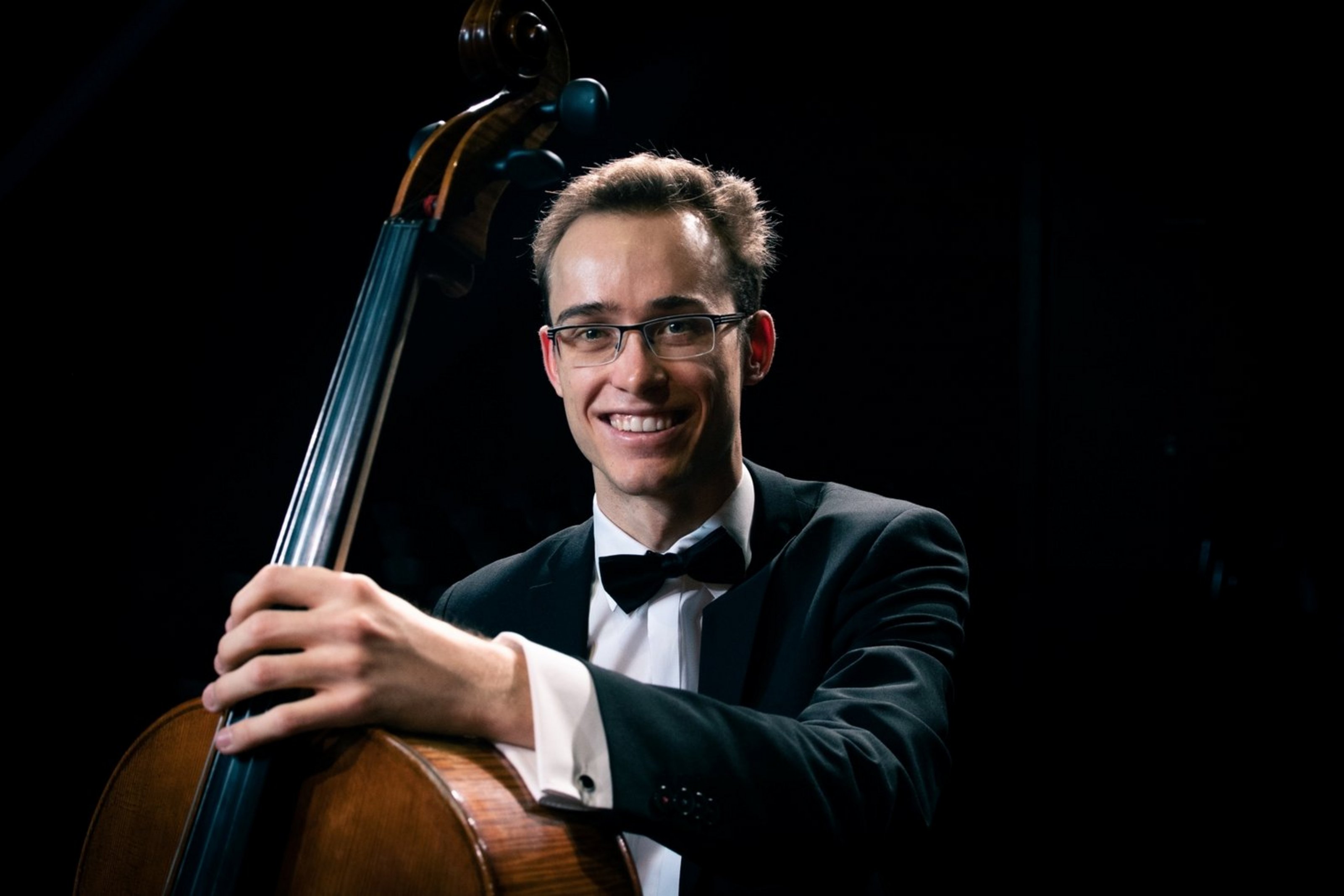 Ein Mann mit Anzug und Brille sitzt hinter einem Cello. Eine Hand hat er auf den Hals des Cellos gelegt.
