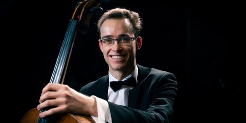 Ein Mann mit Anzug und Brille sitzt hinter einem Cello. Eine Hand hat er auf den Hals des Cellos gelegt.