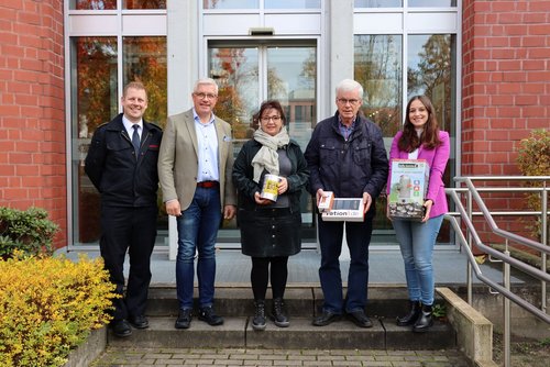 Auf einer Treppenstufe vor einem geklinkerten Gebäude stehen fünf Personen nebeneinander und schauen in die Kamera. Drei Personen halten Gegenstände in den Händen.