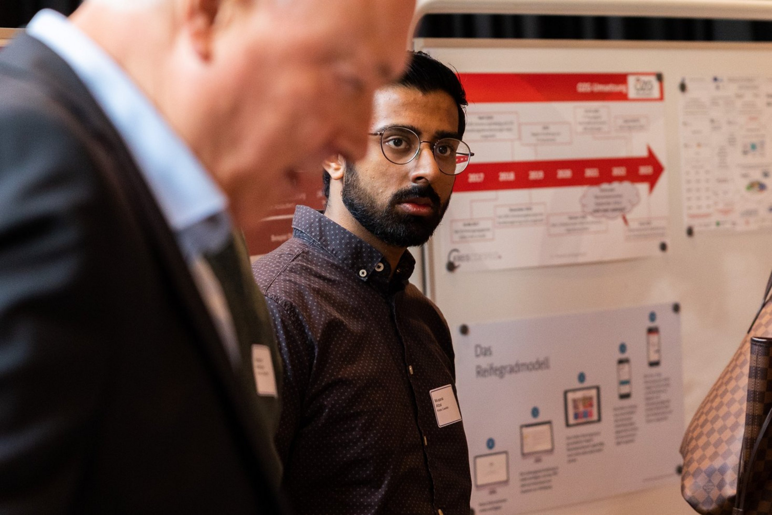 Mann mit Brille vor Wand mit Prozessdiagramm, im Vordergrund unscharfer älterer Mann
