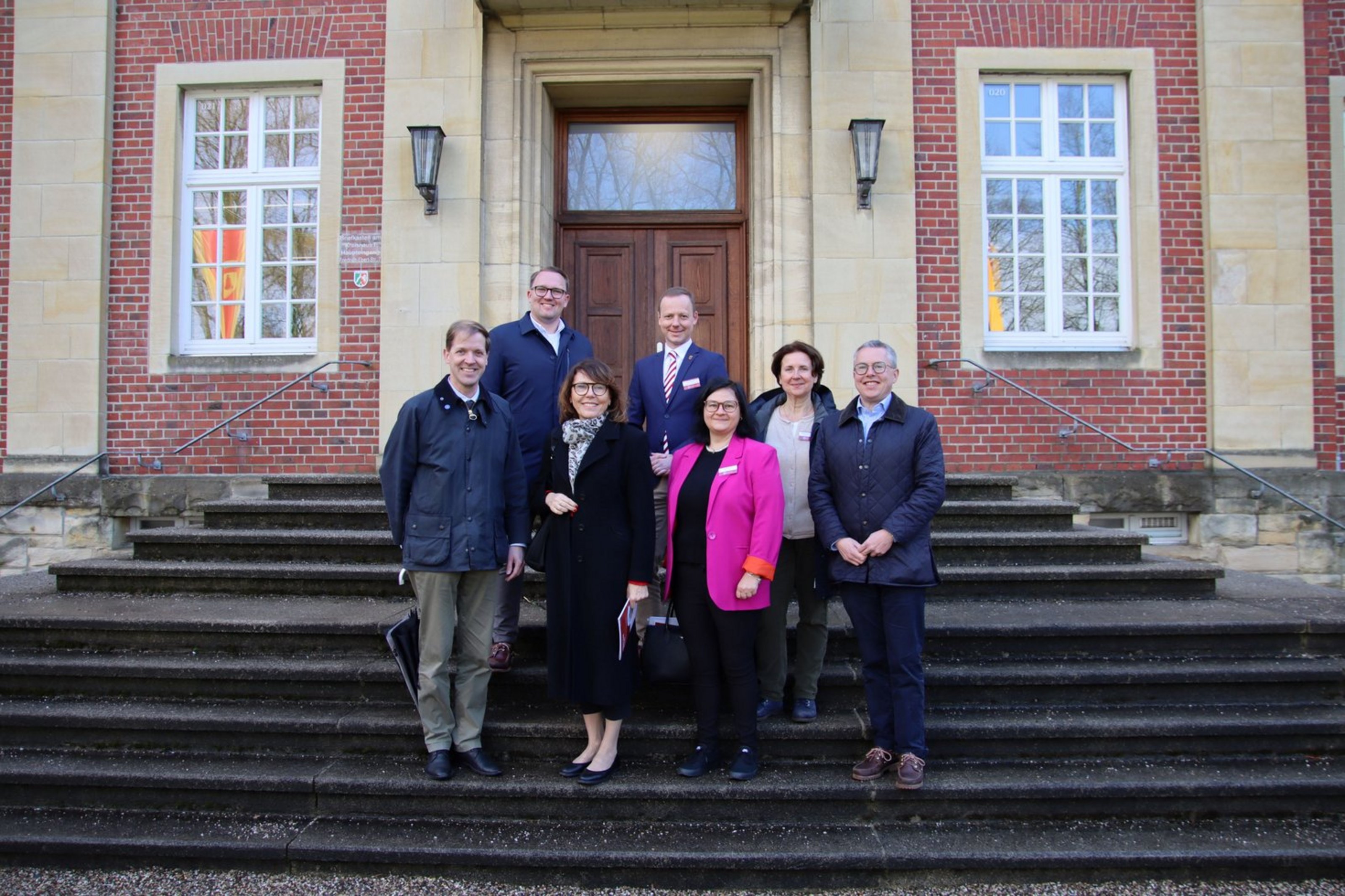 Eine Gruppe mit acht Personen stehen in zwei Reihen auf den Stufen vor dem Eingangstor eines historischen Backsteingebäudes.