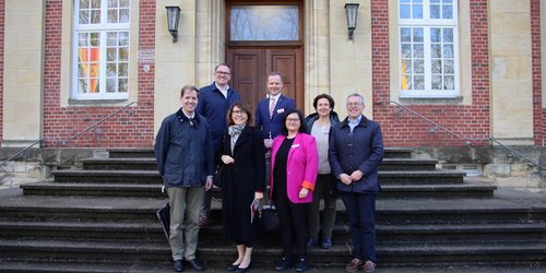 Eine Gruppe mit acht Personen stehen in zwei Reihen auf den Stufen vor dem Eingangstor eines historischen Backsteingebäudes.