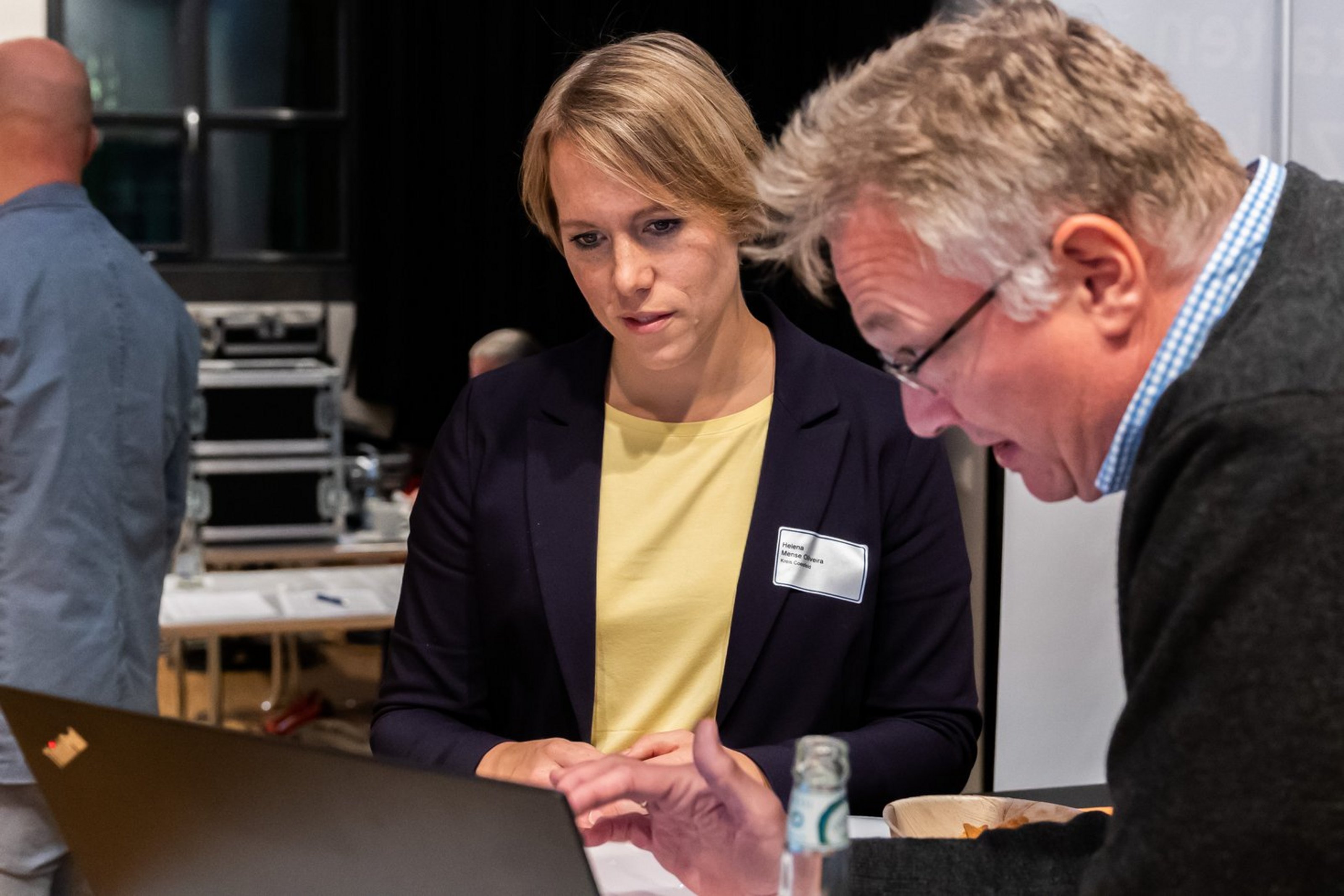 Zwei Personen in einem Innenraum, eine Frau sitzt und schaut auf einen Laptop, ein Mann beugt sich vor und zeigt auf den Laptopbildschirm