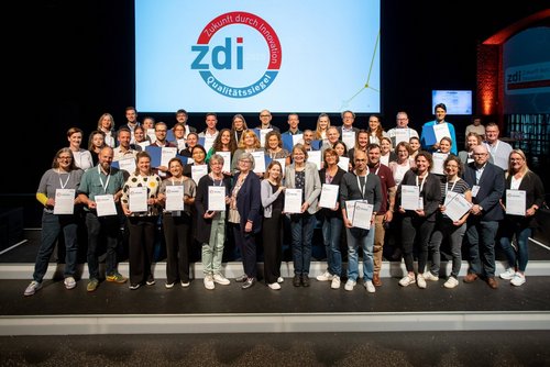 Viele Menschen stehen in drei Reihen auf einer Bühne und präsentieren die Urkunden zum Qualitätssiegel.