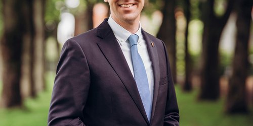 Ein Mann mit Anzug und Krawatte steht auf einem Rasenstück mit Bäumen im Hintergrund. Seine Hände berühren sich vor seinem Bauch. Er lächelt.