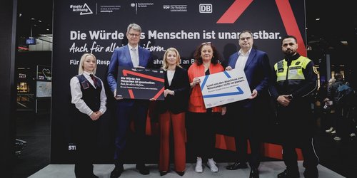 Sechs Personen stehen nebeneinander vor einem großen, schwarzen Plakat  mit der Aufschrift "Die Würde des Menschen ist unantastbar." Der Rest des Schriftzugs ist von den Personen verdeckt. Die Personen halten insgesamt zwei Schilder in der Hand, die den Schriftzug "Für mehr Respekt in Bussen, Bahnen und Bahnhöfen" teilen.