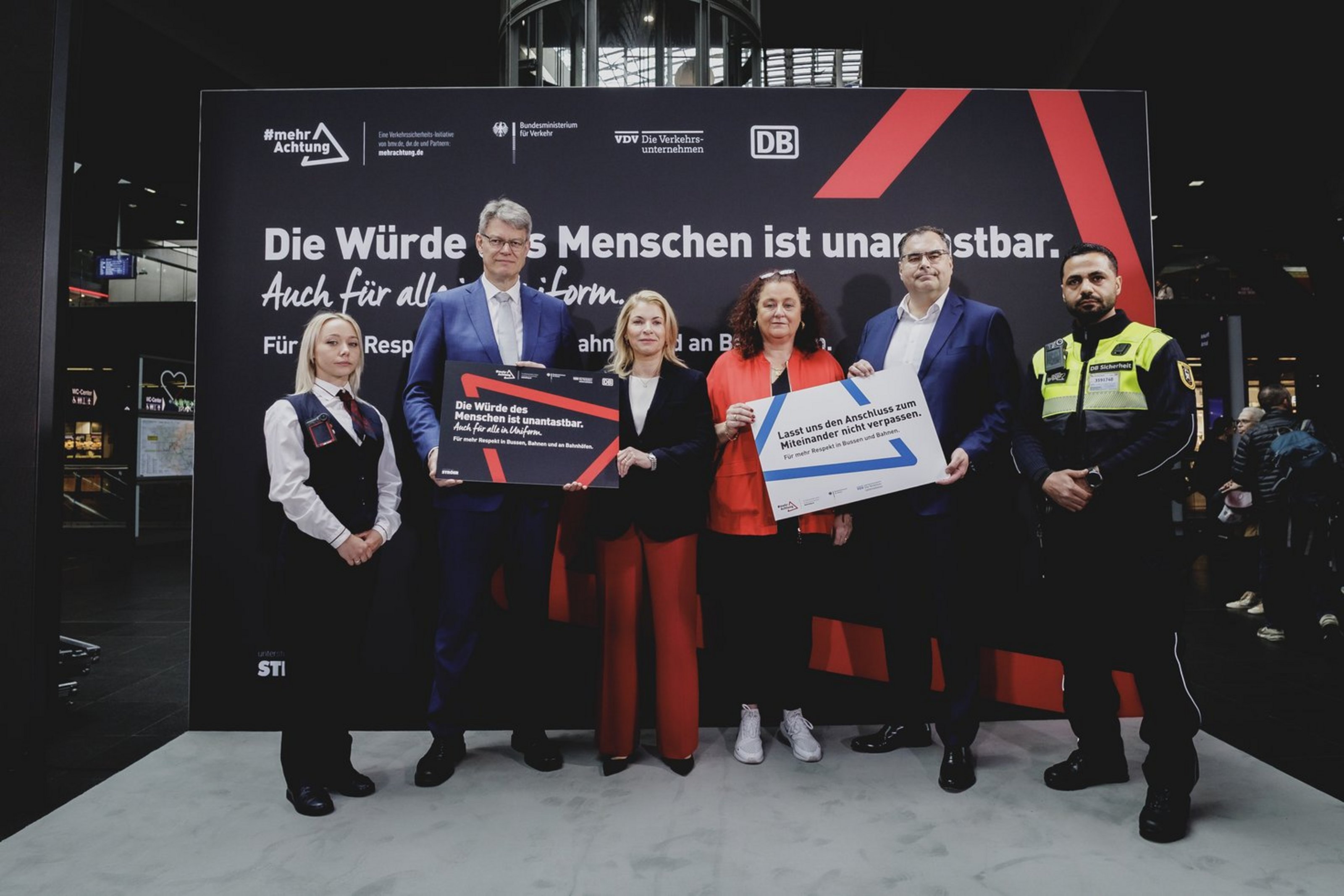 Sechs Personen stehen nebeneinander vor einem großen, schwarzen Plakat  mit der Aufschrift "Die Würde des Menschen ist unantastbar." Der Rest des Schriftzugs ist von den Personen verdeckt. Die Personen halten insgesamt zwei Schilder in der Hand, die den Schriftzug "Für mehr Respekt in Bussen, Bahnen und Bahnhöfen" teilen.