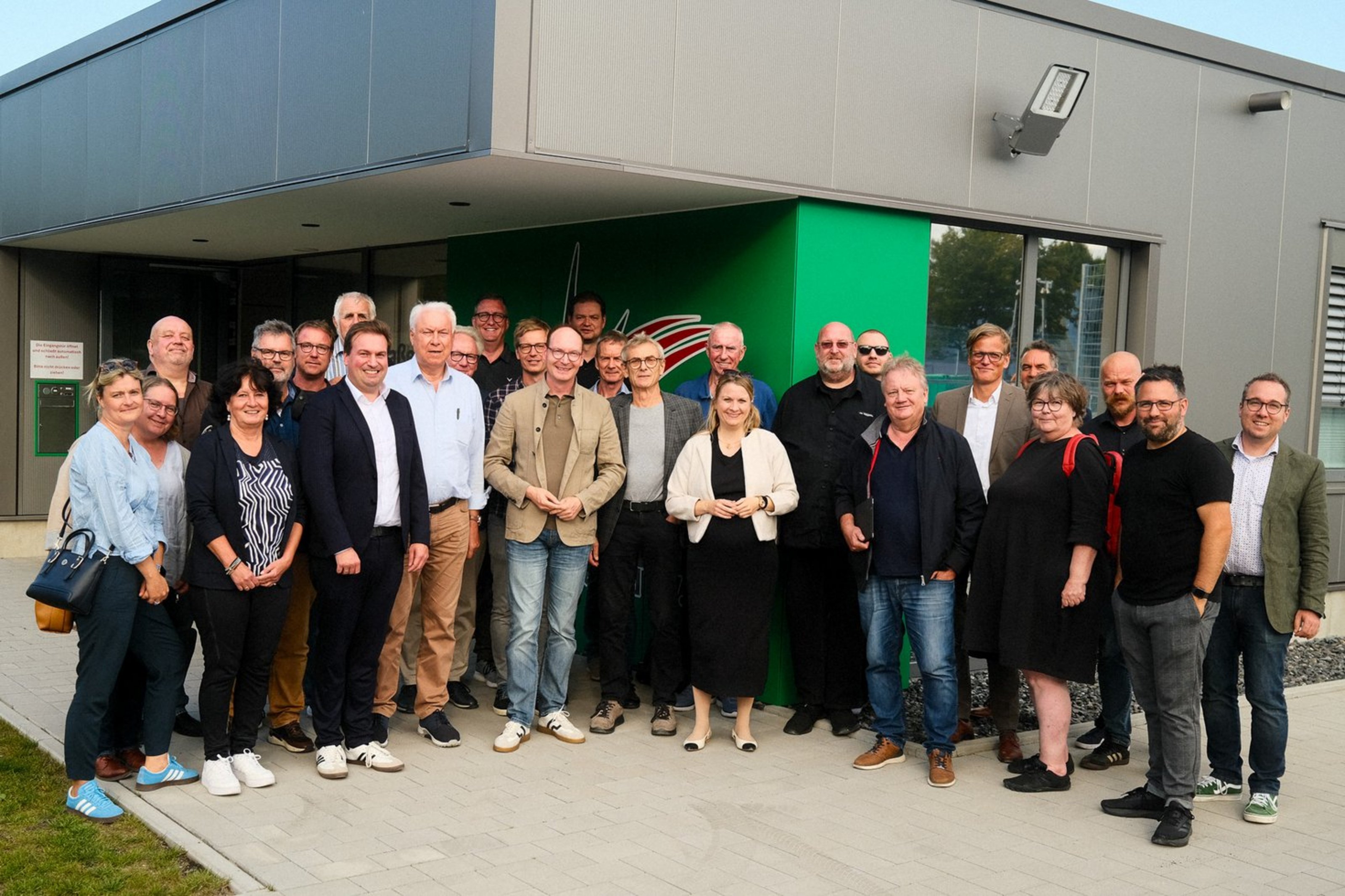 Der Kreisausschuss Kultur, Sport und Ehrenamt vor dem Campus Nord der SG Coesfeld (Foto: Lukas Bertels, Kreis Coesfeld). Eine Gruppe, bestehend aus 27 Personen, steht im Halbkreis vor einem grauen Gebäude mit grünem Eingangsbereich.