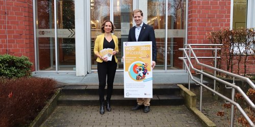 Ein Mann und eine Frau stehen mit Blazer vor einer Eingangstür und halten ein Plakat und einen Flyer in gelb.