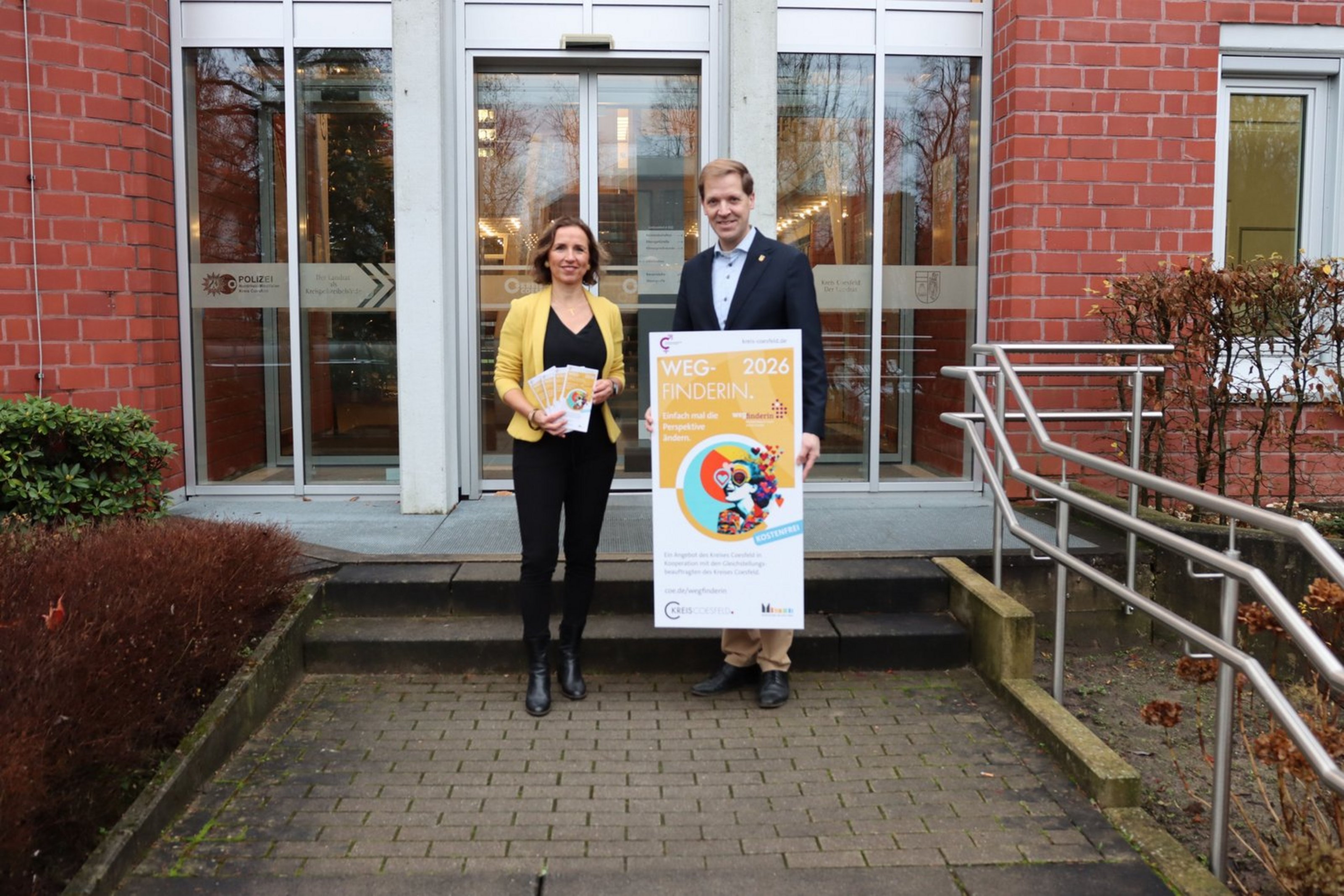 Ein Mann und eine Frau stehen mit Blazer vor einer Eingangstür und halten ein Plakat und einen Flyer in gelb.