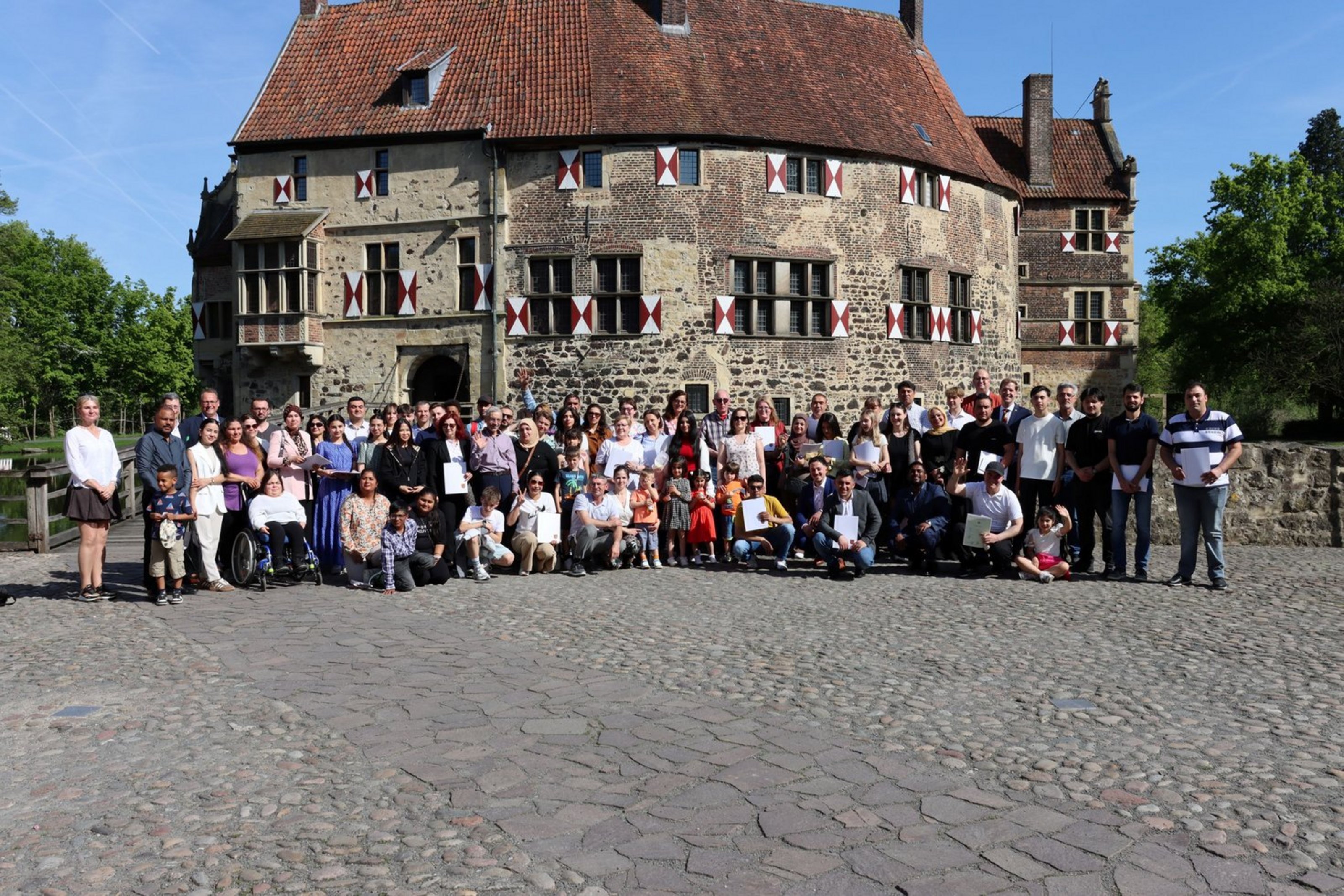 Einbürgerungsfeier am 30. April 2025 auf der Burg Vischering in Lüdinghausen (Foto: Kreis Coesfeld) Eine große Gruppe von Menschen, etwa 80 Personen, steht und sitzt für ein Gruppenfoto vor einer historischen Burg mit Fachwerk und roten Fensterläden. Einige Personen halten Urkunden oder Papiere in den Händen. Im Vordergrund sind auch Kinder und eine Person im Rollstuhl zu sehen. Die Gruppe steht auf einem Pflastersteinplatz bei sonnigem Wetter. Im Hintergrund ist die Burg mit einem roten Ziegeldach, vielen Fenstern und einer hölzernen Zugbrücke zu sehen. Links und rechts sind Bäume und blauer Himmel.