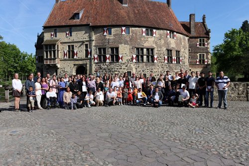 Eine große Gruppe von Menschen, etwa 80 Personen, steht und sitzt für ein Gruppenfoto vor einer historischen Burg mit Fachwerk und roten Fensterläden. Einige Personen halten Urkunden oder Papiere in den Händen. Im Vordergrund sind auch Kinder und eine Person im Rollstuhl zu sehen. Die Gruppe steht auf einem Pflastersteinplatz bei sonnigem Wetter. Im Hintergrund ist die Burg mit einem roten Ziegeldach, vielen Fenstern und einer hölzernen Zugbrücke zu sehen. Links und rechts sind Bäume und blauer Himmel.