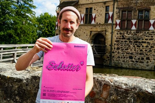 Ein Mann hält das Plakat zu Baddabäm in pinker Gestaltung vor sich.