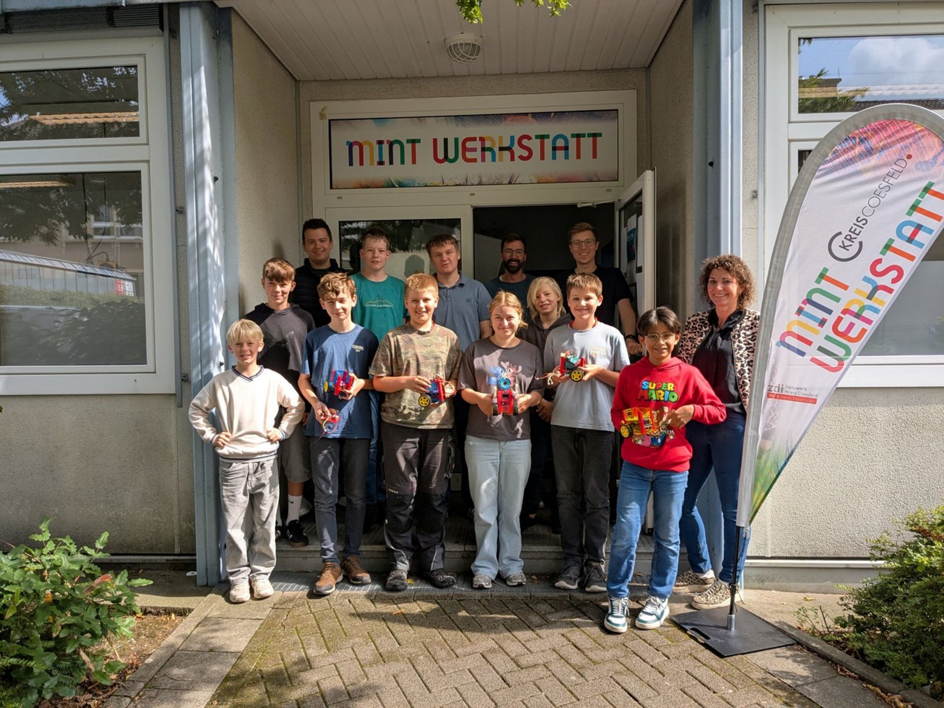 Eine Gruppe junger Teenager und einige Erwachsene stehen in einem Hauseingang. Über der Tür steht groß Mint Werkstatt.
