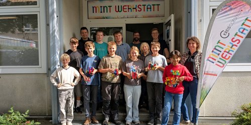 Eine Gruppe junger Teenager und einige Erwachsene stehen in einem Hauseingang. Über der Tür steht groß Mint Werkstatt.