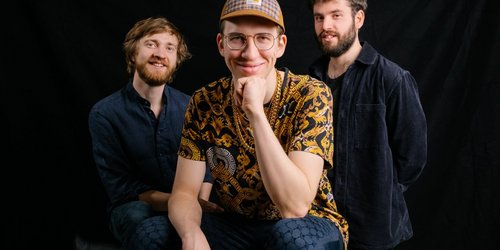 Das Nils Kugelmann Trio (Foto: Uli Neumann-Cosel) Drei Männer sind vor schwarzem Hintergrund zu sehen. Einer von ihnen kniet vorne, die anderen stehen rechts und links hinter ihm.