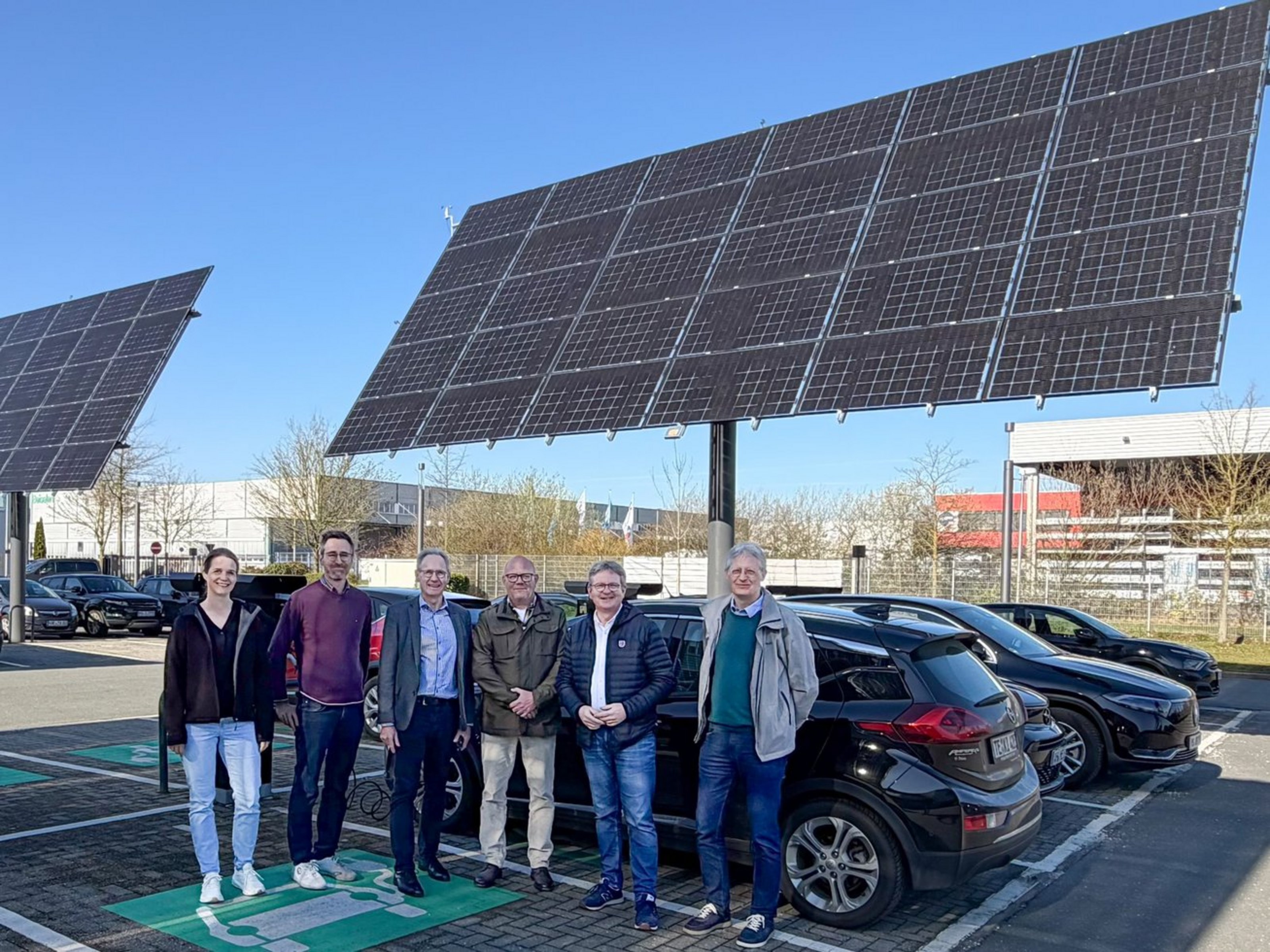 Sechs Personen stehen vor einem Parkplatz, auf dem große Solarmodule stehen.