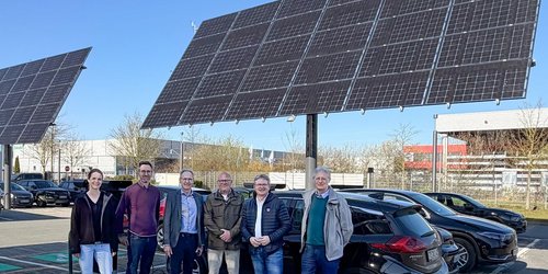 Sechs Personen stehen vor einem Parkplatz, auf dem große Solarmodule stehen.