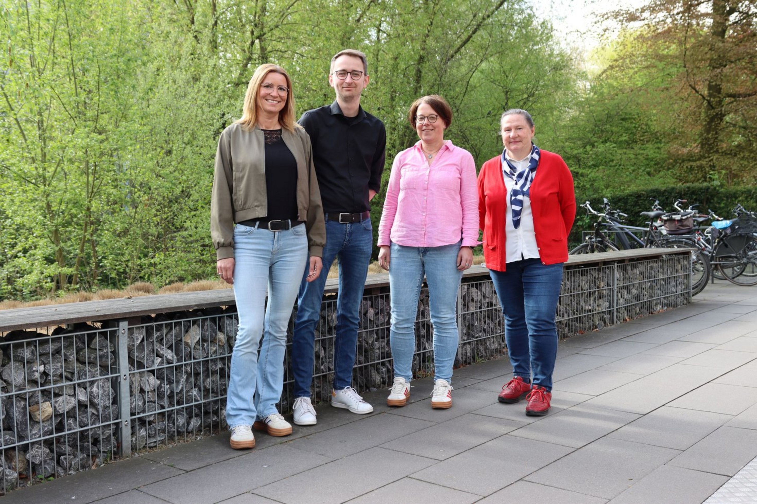 Vier Personen stehen nebeneinander vor einer Mauer aus Steinen.  Es sind drei Frauen und ein Mann. Alle lächeln in die Kamera.