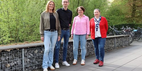 Vier Personen stehen nebeneinander vor einer Mauer aus Steinen.  Es sind drei Frauen und ein Mann. Alle lächeln in die Kamera.